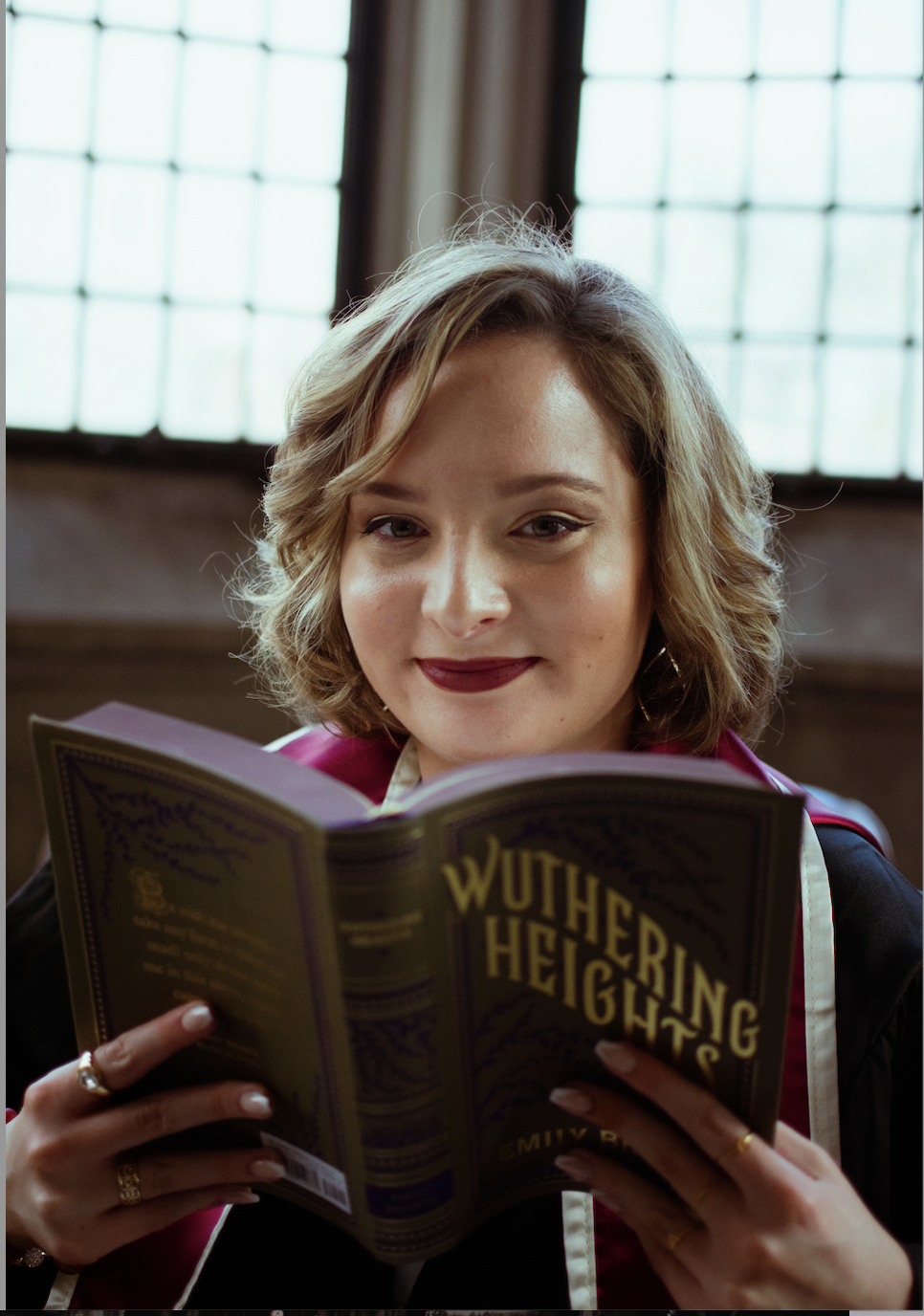 Photograph of Emily, wearing graduation robes and holding up a vintage copy of Wuthering Heights.
