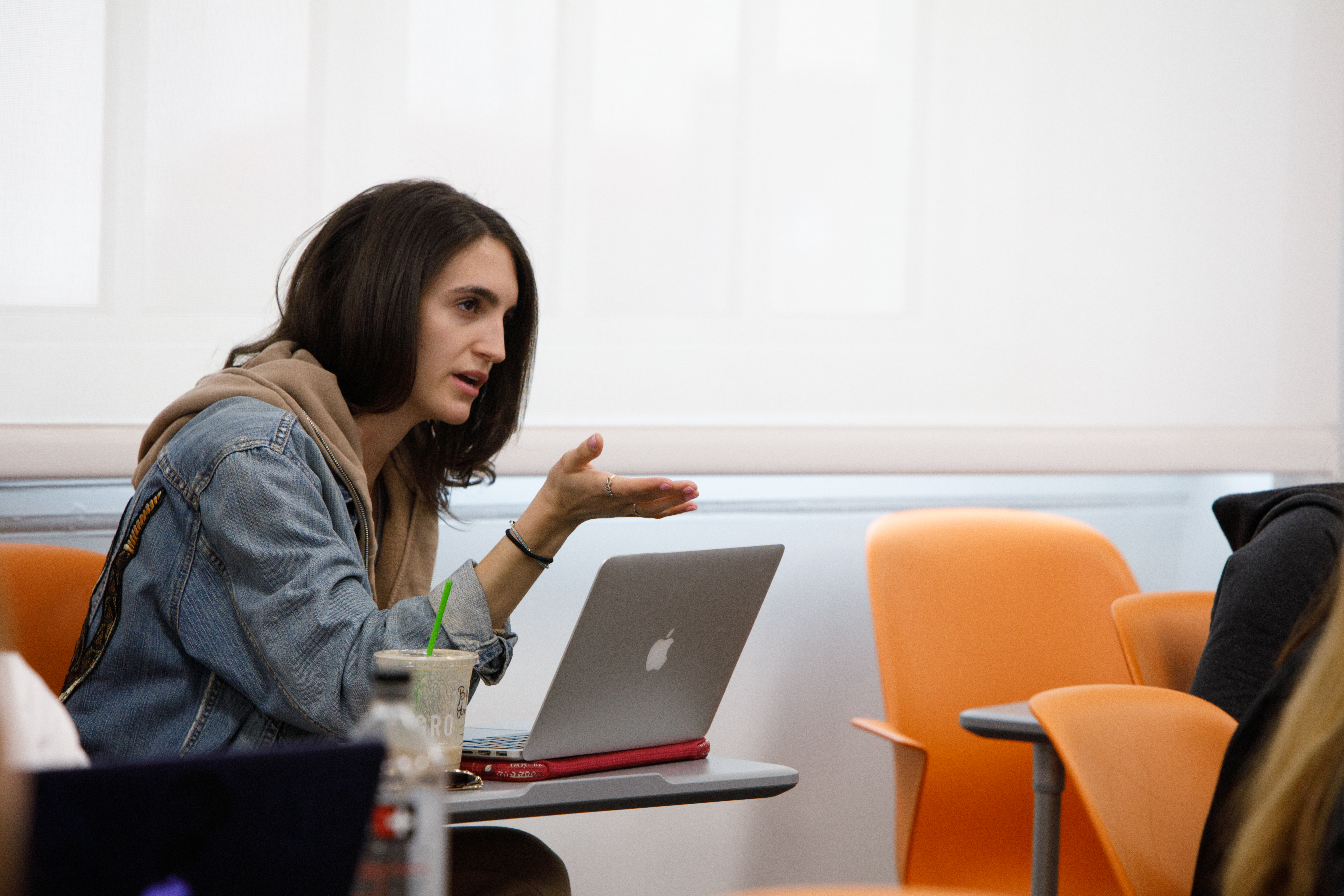 A young woman sits at a desk with a computer in front of her, speaking to her classmates