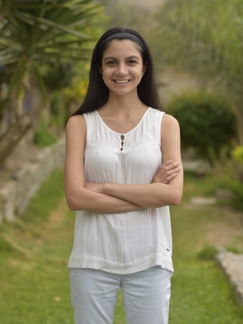 Photograph of Ana, wearing a white top and light blue pants stands on a grass path, smiling, surrounded by greenery.