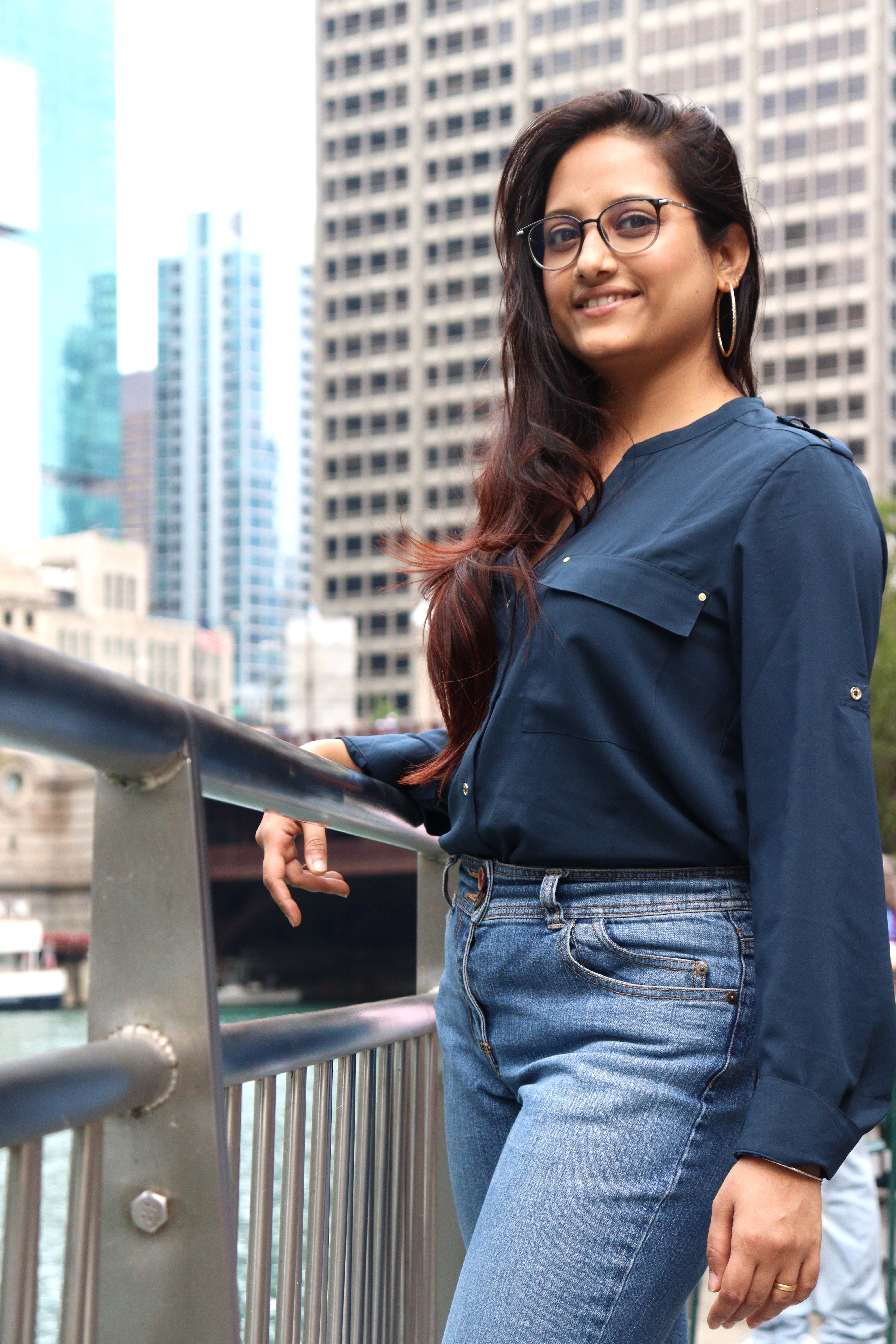 Photograph of Priyanka, wearing jeans and a blue blouse, standing in front of a river in an urban setting, with her arm over a railing.