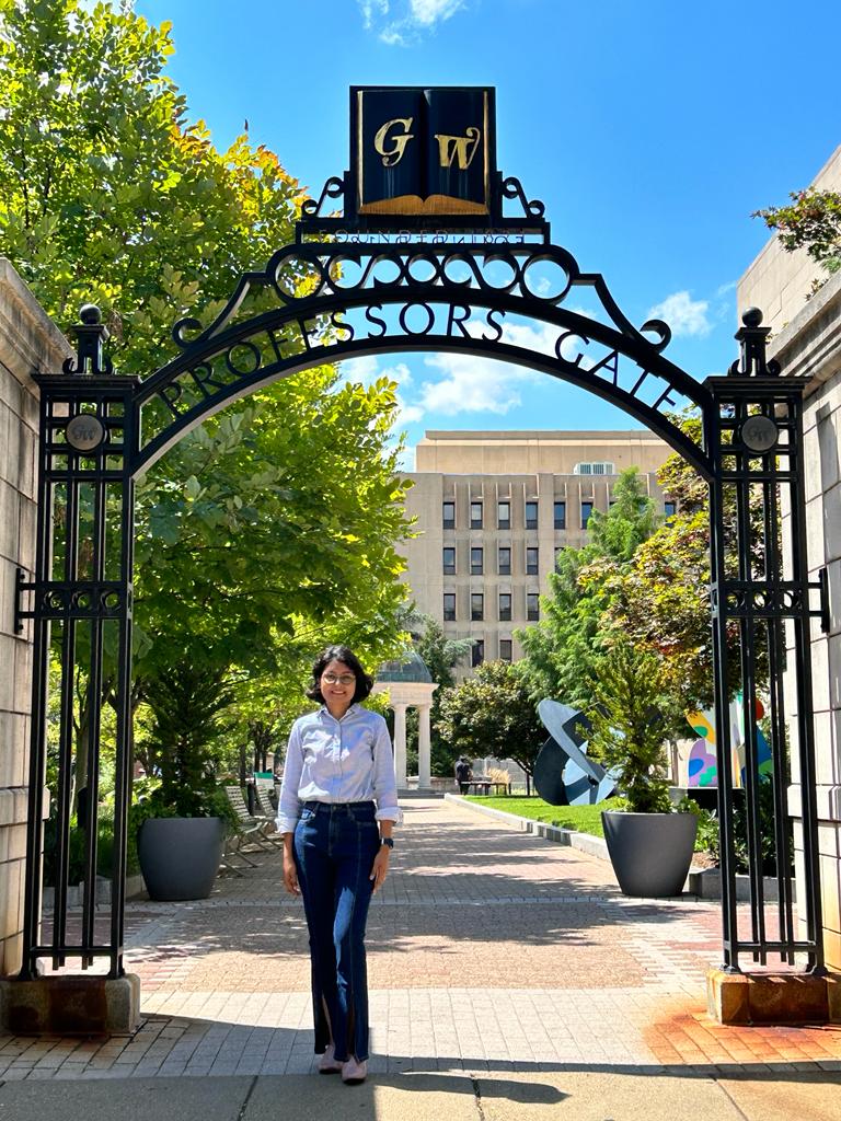 Photograph of Soumi standing under the George Washington University gate, under a blue sky and in front of green trees.