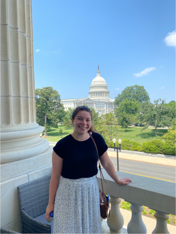Photograph of Isabelle, wearing a shirt and skirt, standing on a balcony with the Capitol building in the background.
