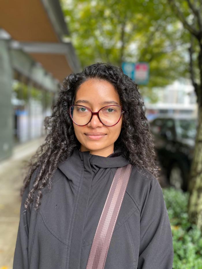 Photograph of Leenu, wearing glasses, a black jacket, and a cross-body bag. In the background, there are shops and trees.