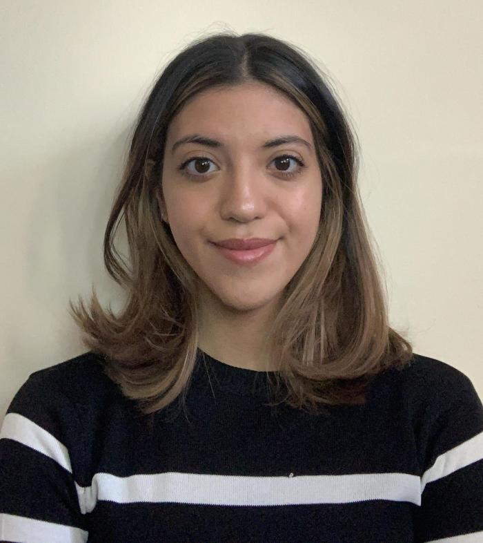 Photograph of Madi, wearing a striped shirt and standing against a plain wall.