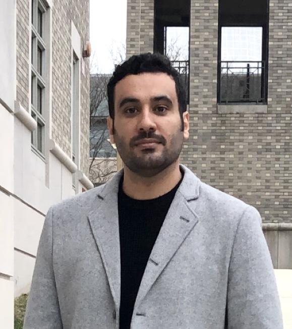 Photograph of Raad, standing outside in front of some buildings, wearing a gray coat and black shirt.