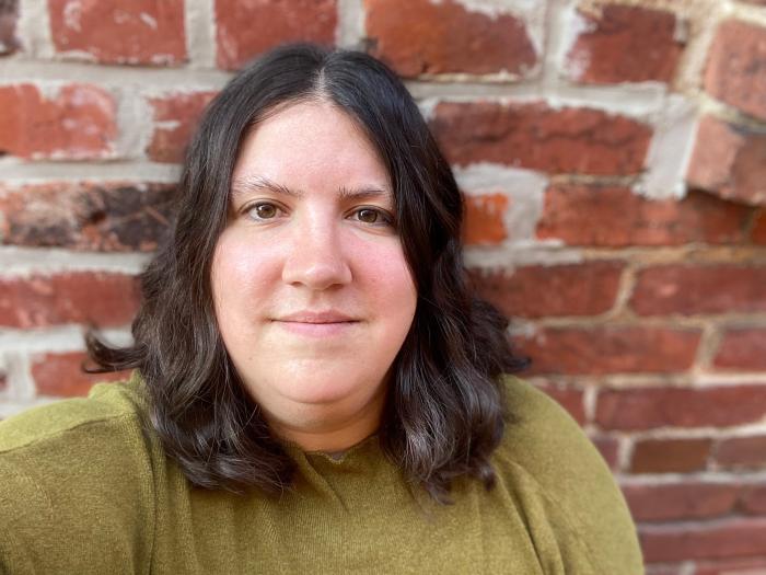 Photograph of Rachel, wearing a green shirt and standing in front of a brick wall.